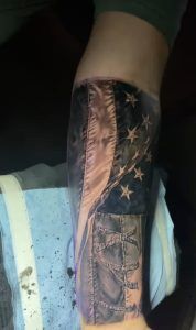 Tattoo on a forearm depicting a realistic, weathered American flag with visible stitching details and frayed edges. The flag appears to be waving, with a dark blue field of stars at the top and vertical red and white stripes extending downward. The shading creates a worn, fabric-like texture for a three-dimensional effect.