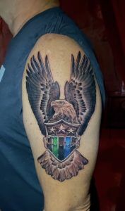 Tattoo on an upper arm showing a realistic bald eagle with wings spread high, clutching a shield in its talons. The shield features three white stars on the top section and vertical stripes in green, black, blue, and red on the lower section. The eagle’s feathers are shaded in black and gray, creating a three-dimensional effect.