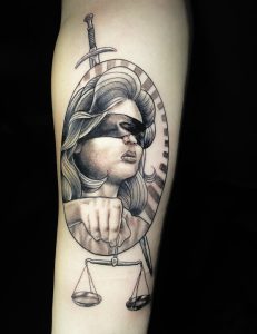 Tattoo on a forearm depicting Lady Justice with long flowing hair and a blindfold over her eyes. She holds a set of balanced scales in her hand extending below the oval frame. A sword is positioned vertically behind her head, pointing upward. The design uses fine line work and shading to create depth and a classical illustration style.