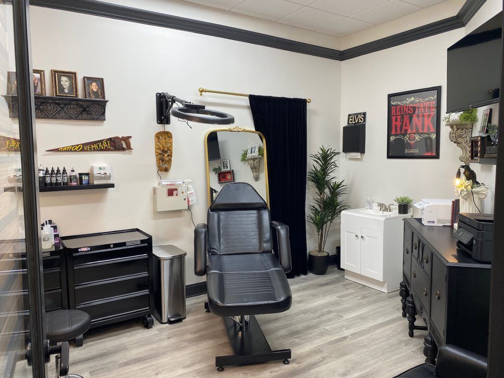 A clean, stylish tattoo studio room at Jason Elvis Martin Studio in Hendersonville, Tennessee. The space features a black leather tattoo chair centered under a circular LED light, with a black curtain, ornate gold-framed mirror, and wall-mounted décor. Art and vintage-style signs—including one reading “Tattoo Removal” and another saying “Reinstate Hank”—decorate the white walls. A black tool chest, white sink cabinet, and black vintage dresser with a printer and small plants contribute to the studio’s professional yet artistic vibe.